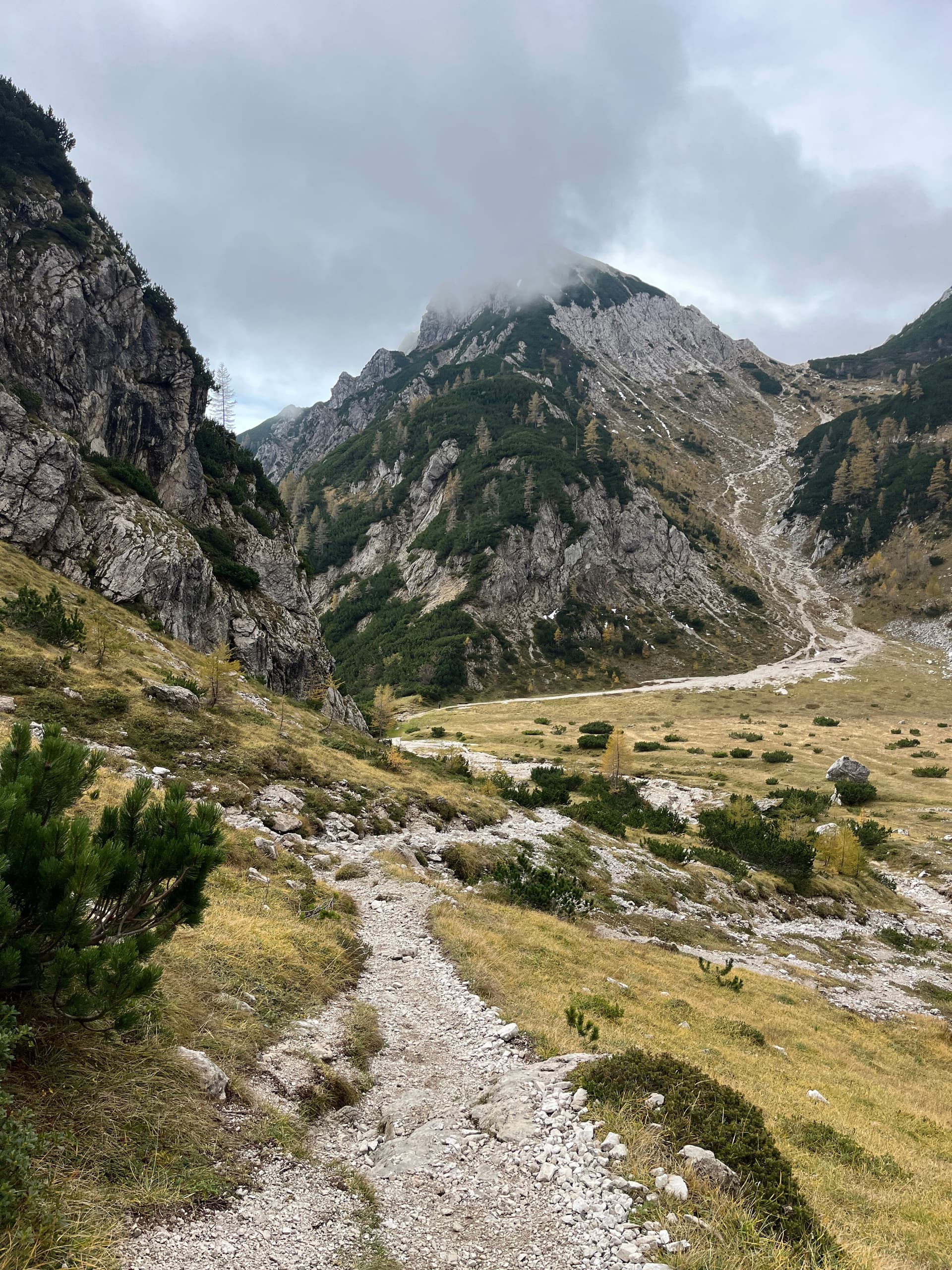 Rocky hiking trail ascending through a grassy mountain valley under a cloudy sky.