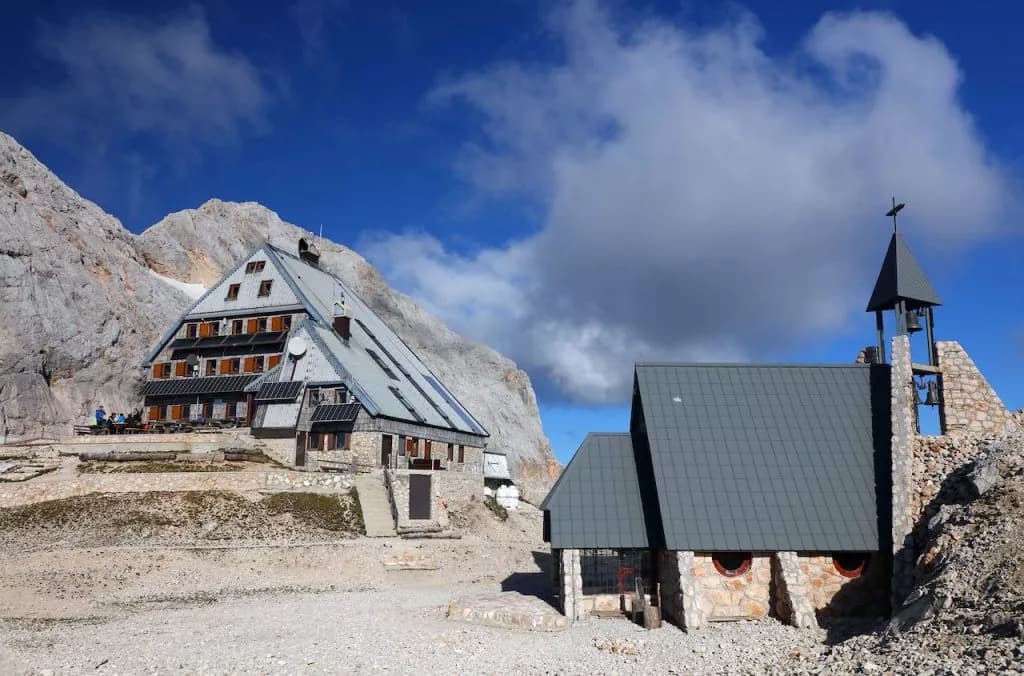 Triglav lodge Kredarica and small stone chapel nestled against a large rocky mountain under a blue sky.