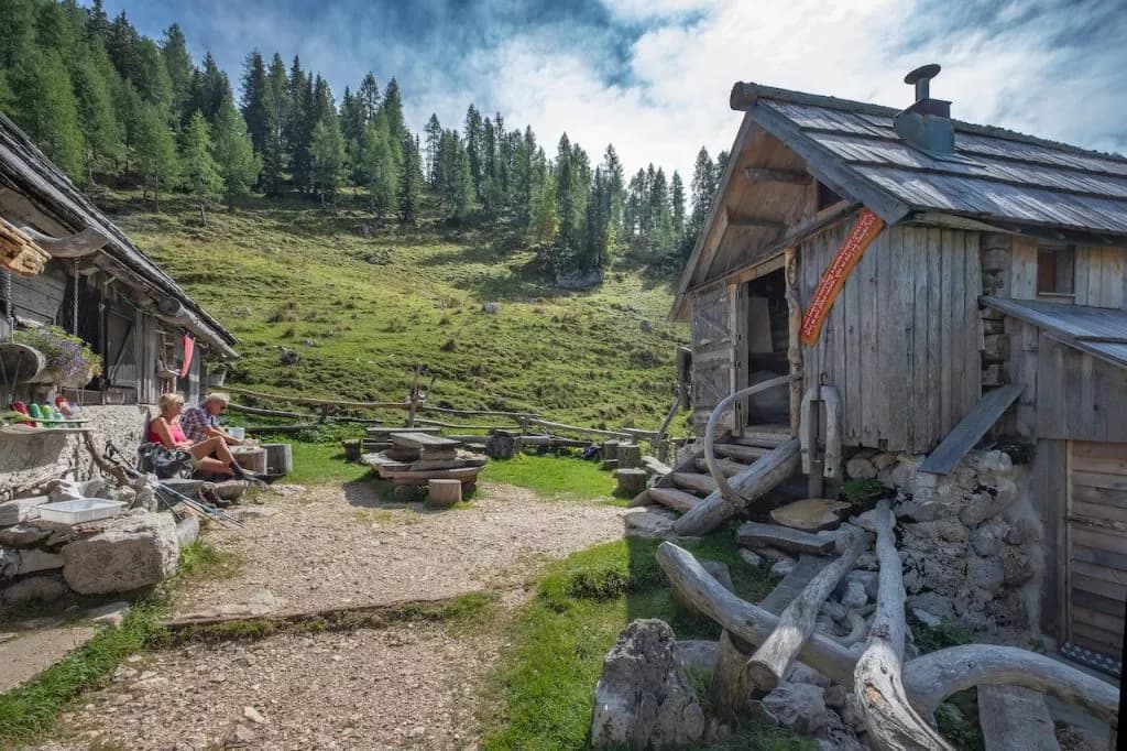 Hikers resting outside rustic wooden shelter on grassy slope at Bregar shelter on Viševnik pasture.