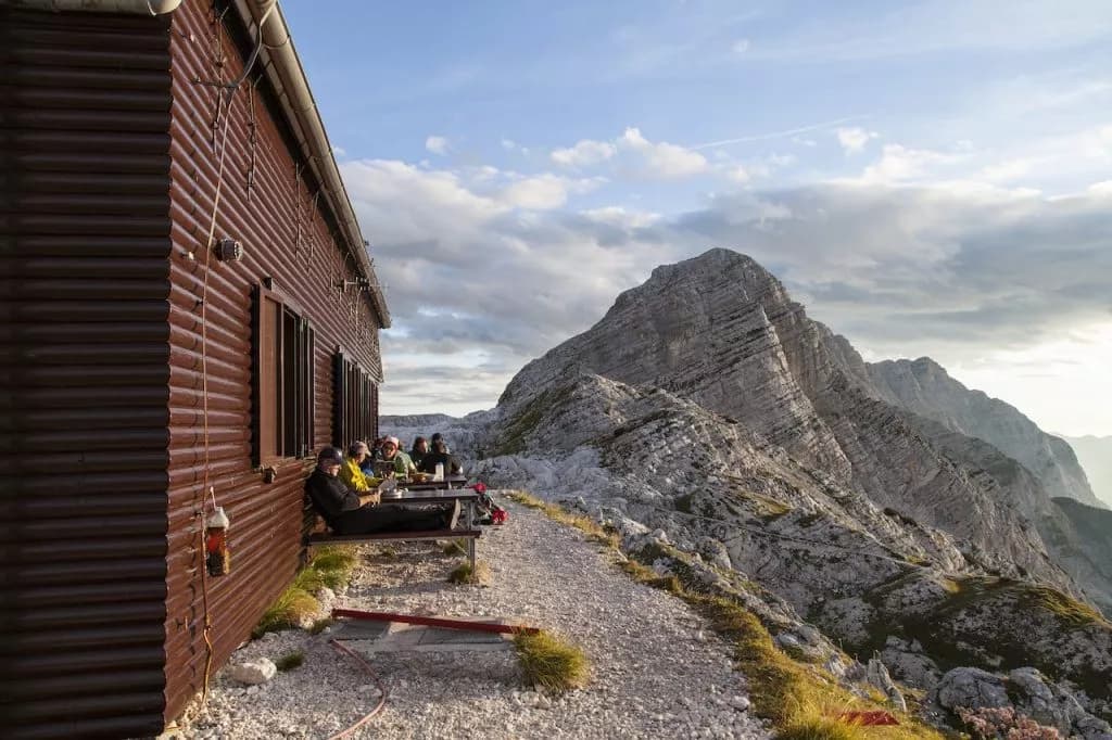 Hikers resting outside Zasavska hut at Prehodavci with massive layered mountain backdrop.