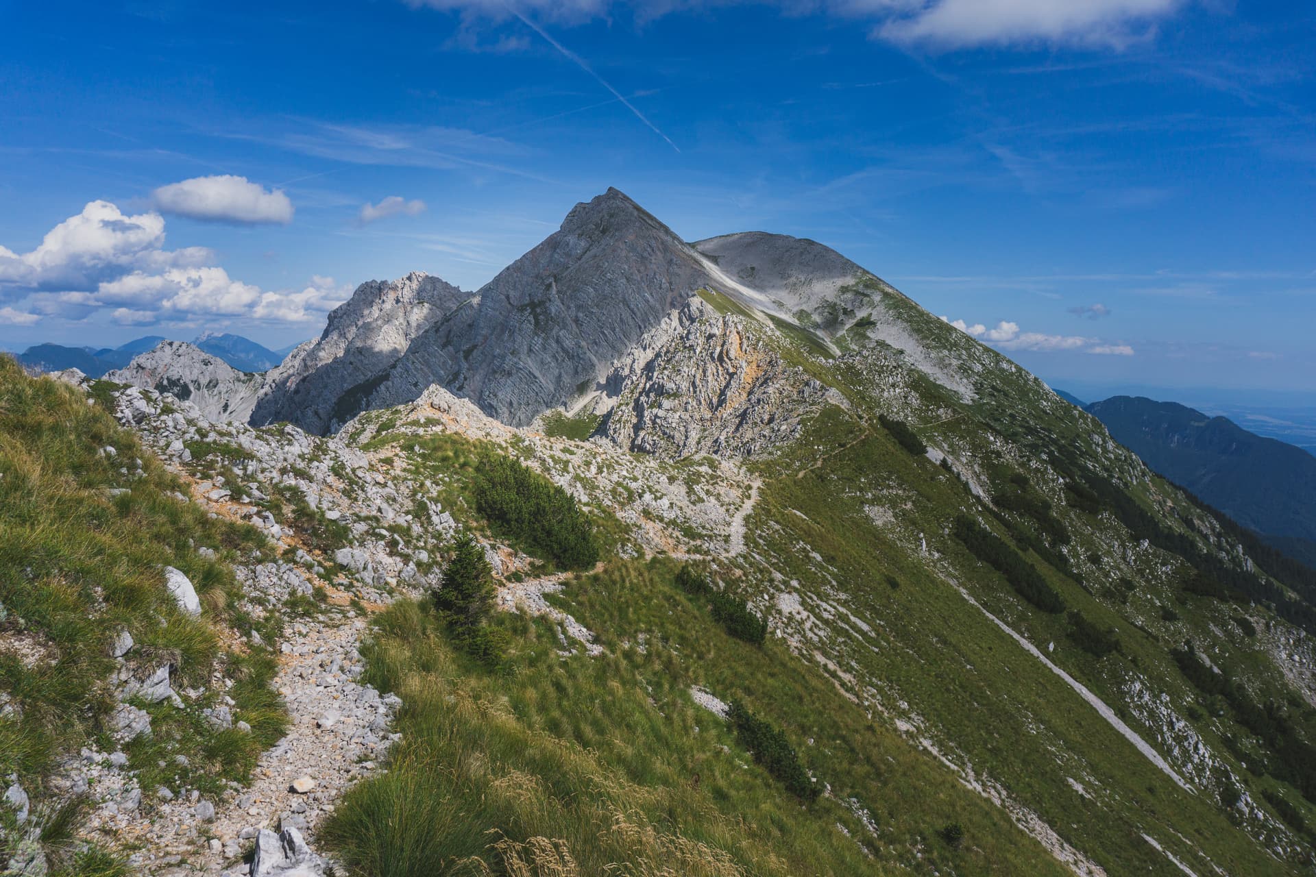 Hiking trail along grassy ridge toward rocky mountain peak under blue sky