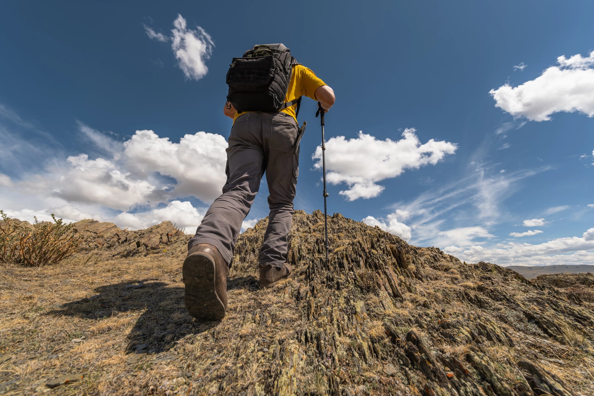 Een mannelijke wandelaar met een rugzak wandelt steil omhoog op een rotsachtige helling, waarbij hij opzettelijk de agressieve loop van zijn wandelschoensole blootstelt, vastgelegd vanuit een laag perspectief onder een zonnige lucht.