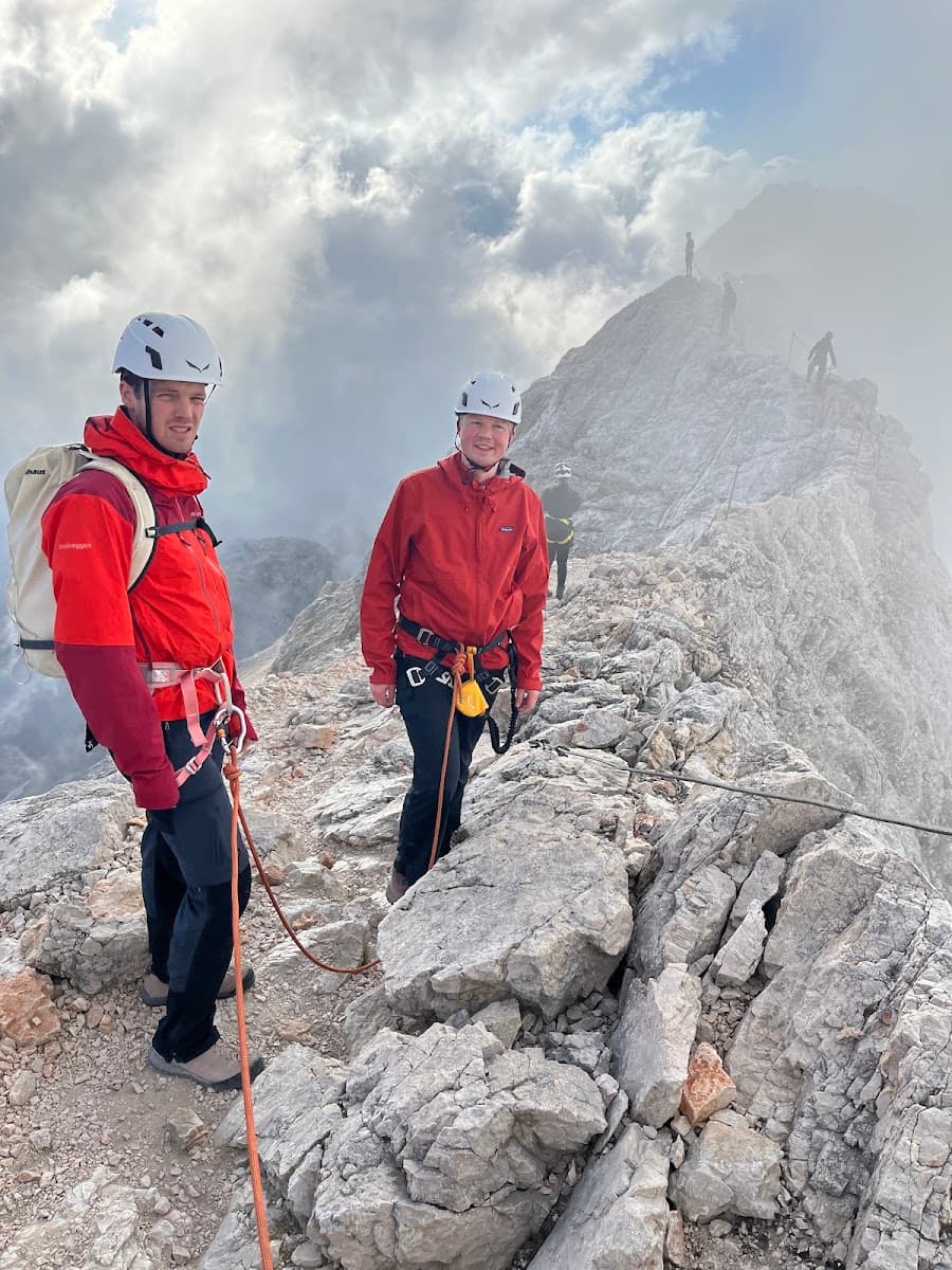 Climbers in red jackets and helmets hiking on a rocky mountain ridge with clouds