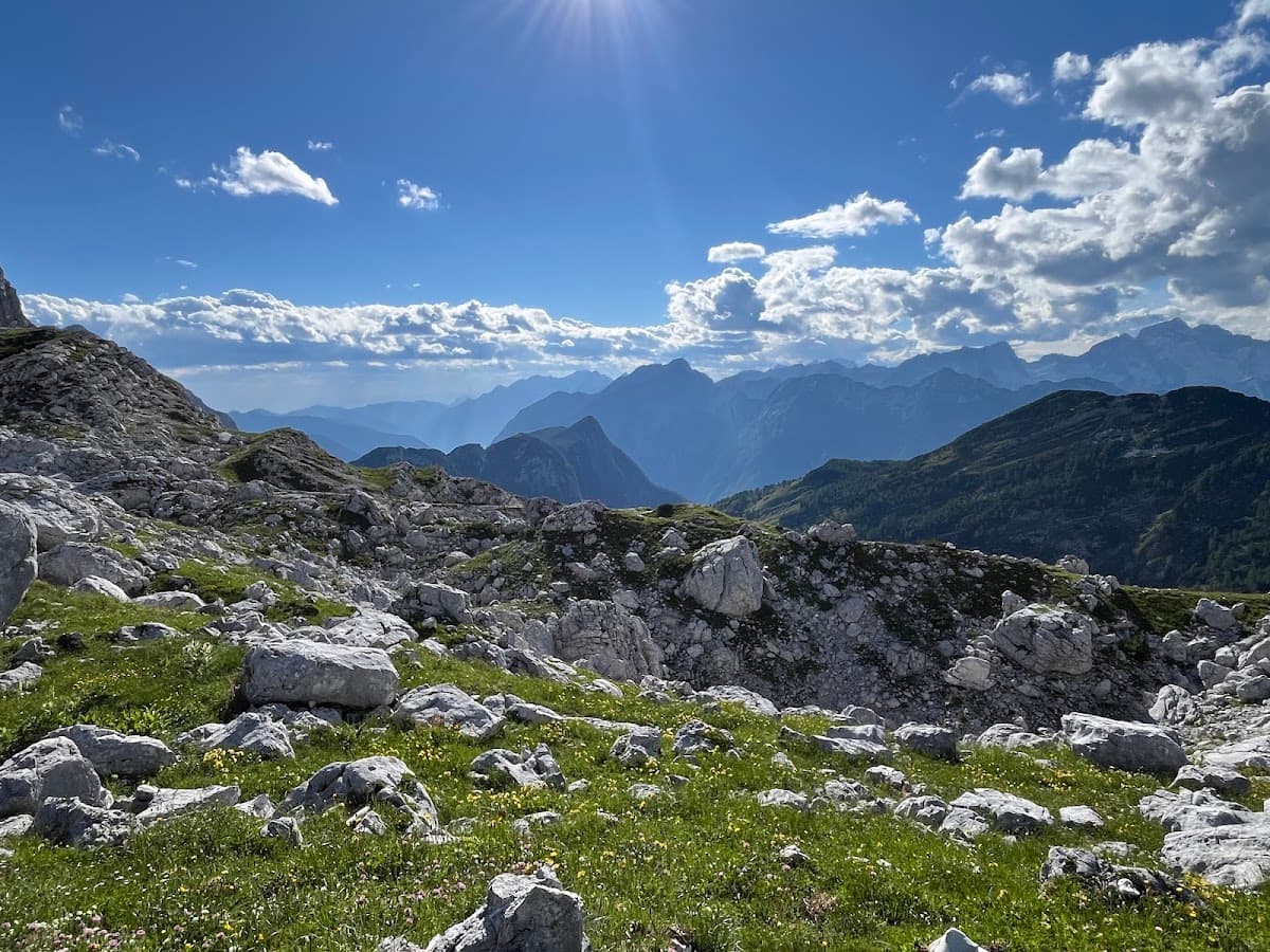 Rocky alpine landscape with green grass, distant mountains, and blue sky with clouds.