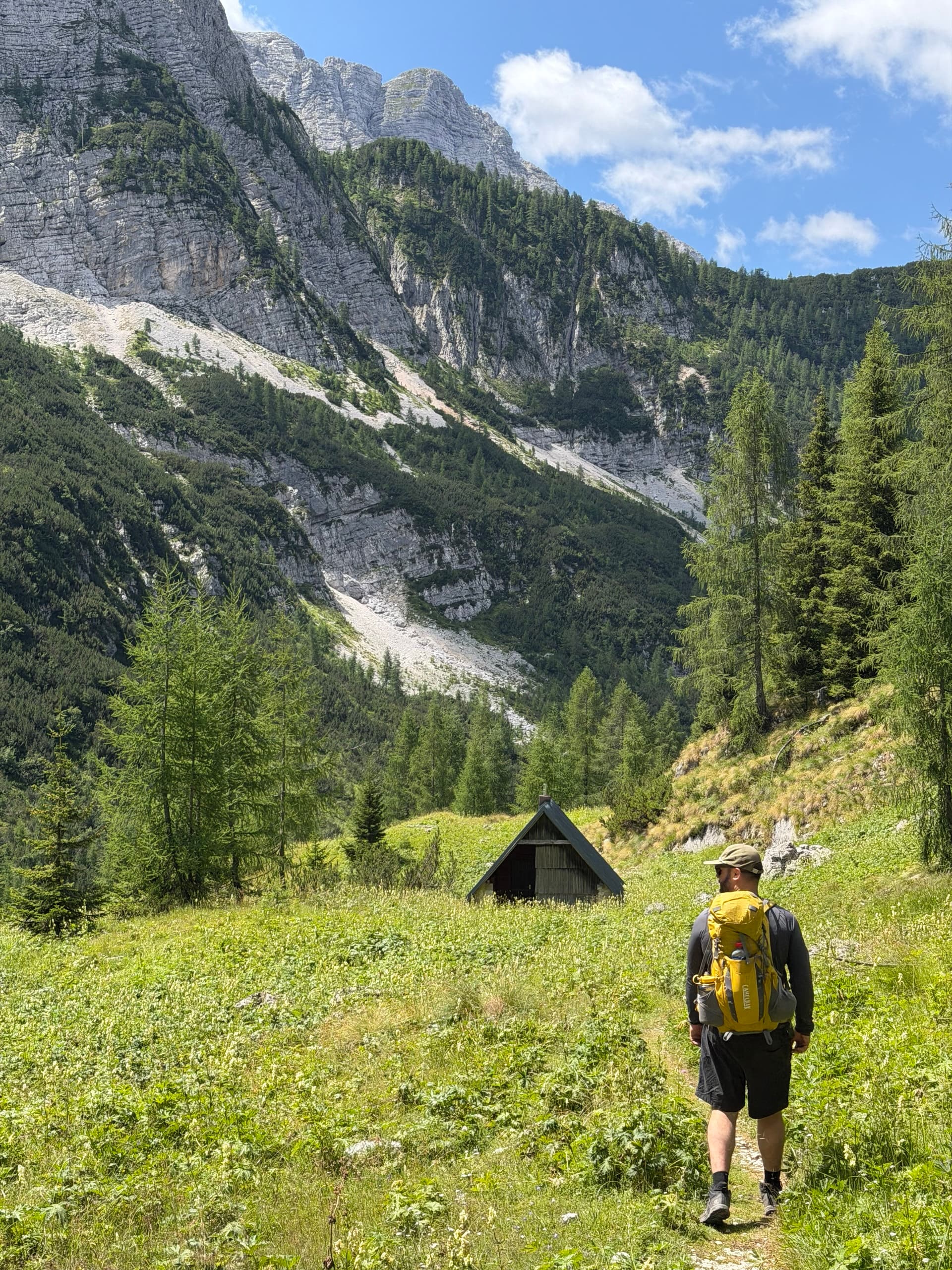Hiker with yellow backpack walking across alpine meadow toward small wooden shepherd hut with rocky mountain backdrop