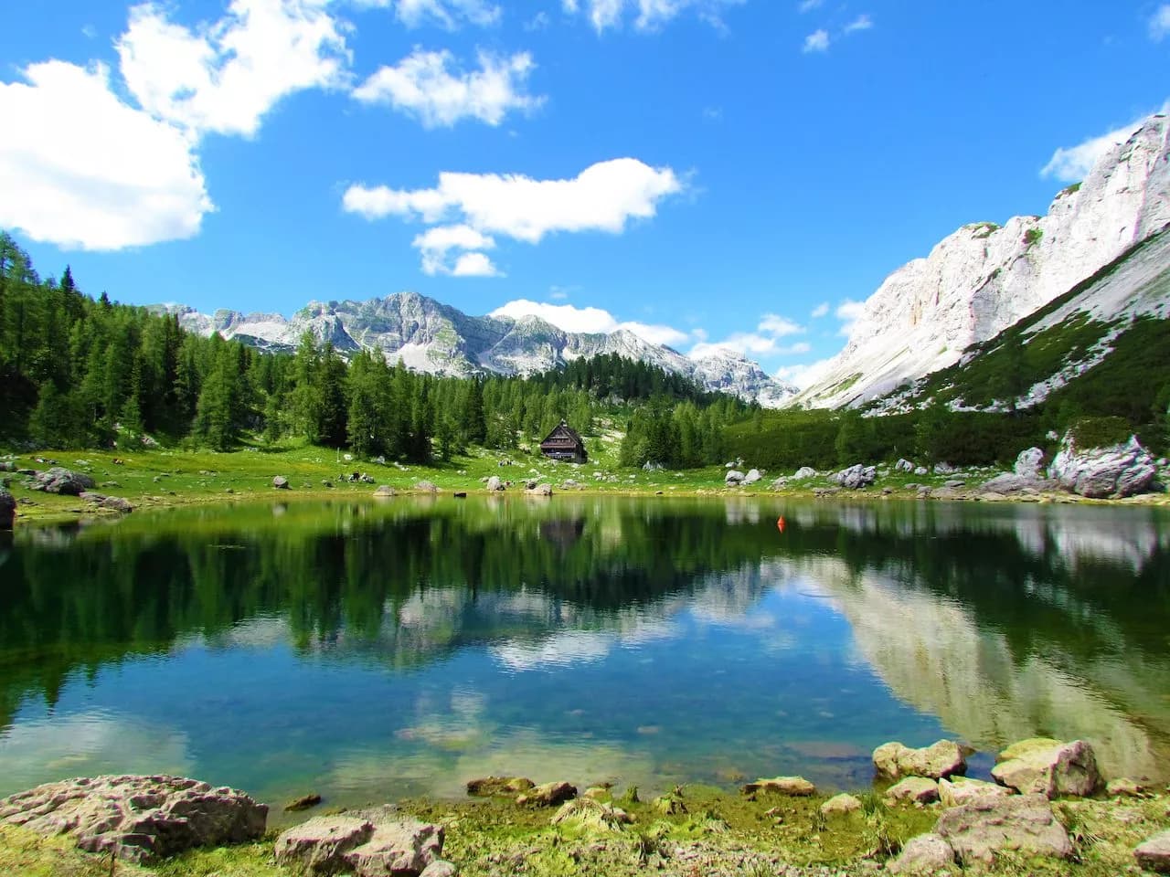 Alpine lake reflecting mountains and forest under a blue sky with white clouds, small lodge visible.